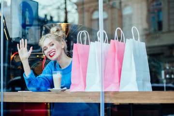 Charming blond hair female say hello while drinking coffee and relaxing after good shopping. Photo taken through the window.