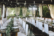 Decorated Elegant Wooden Wedding Table For Banquet Outdoor In Garden Gazebo With Lamp, In The Style Of Rustic With Eucalyptus And Flowers, Porcelain Plates, Glasses, White Chairs