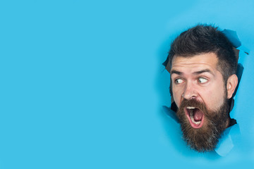 Wall Mural - View of male face through hole in blue paper. Surprised bearded man making hole in paper. Cute attractive man looking through hole. Copy space for advertising, to insert text or slogan. Discount, sale