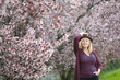 © IrinaN - Caucasian woman with long blond hair and purple fedora hat near blossoming tree, one hand up holding hat, second hand in pocket