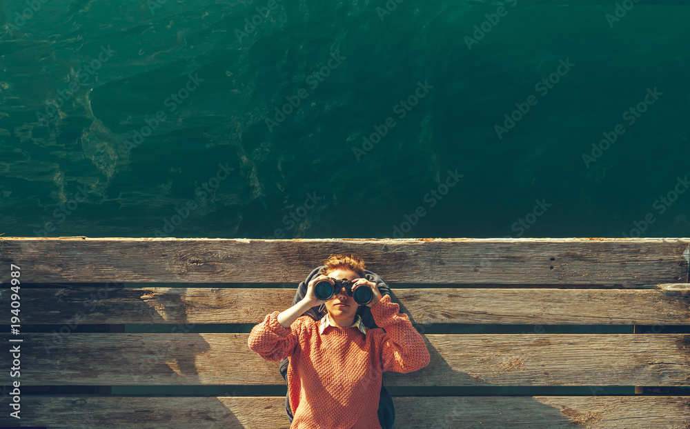 Young Beautiful Girl Lies On A Pier Near The Sea And Looks Through Binoculars On The Sky. Travel Search Journey Concept - obrazy, fototapety, plakaty 