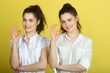 © Alexandr - two beautiful joyful sisters twin girls in white blouses on a yellow background showing gestures of hands a sign of success
