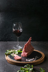 Grilled sliced rack of lamb with yogurt mint sauce served with green salad young beetroot leaves, glass of red wine, pink salt on round wooden slate board over dark black table background. Copy space