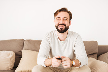 Sticker - Photo of happy guy 30s having beard and mustache resting at home on couch, and watching TV with remote control in hands