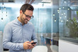 © Bojan - Smiling cheerful young man in smart casual wear holding smart phone and looking at it while standing in modern office.