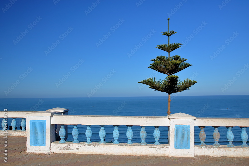 Piękny widok na morze, na pierwszym planie biało-niebieska ozdobna balustrada, po prawo nieduże zielone drzewko, balustrada w style andaluzyjskiej architektury, błękitne niebo - obrazy, fototapety, plakaty 
