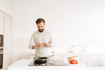 Sticker - Photo of european man with short brown hair and beard cooking omelet with tomato in kitchen, while having dinner at home