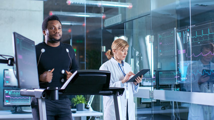 Wall Mural - male athlete runs on a treadmill with electrodes attached to his body while sport scientist holds ta