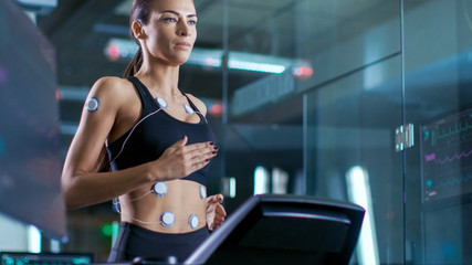 Wall Mural - beautiful woman athlete runs on a treadmill with electrodes attached to her. in the background labor