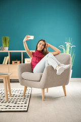 Poster - Beautiful young woman taking selfie while sitting in comfortable armchair at home