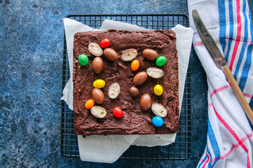 Dark chocolate brownie square on a wire rack decorated with creamy chocolate eggs. Blue stone background.