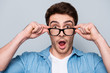 Close up portrait of astonished, shocked, attractive guy holding eyelets of glasses on his face with fingers, having wide open mouth, looking out spectacles, isolated on grey background