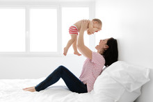 Smiling Mother Sitting On Bed Lifting Baby In The Air