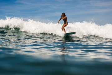 Beautiful young indonesian woman in bikini surfing wave in Bali on the background of blue sky, clouds and tropical beach