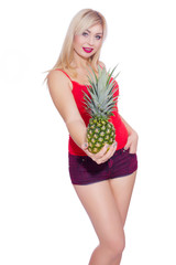 A girl in a red T-shirt and red denim shorts holds a pineapple in one hand and holds it to the camera. A woman holds a second hand in her pocket. Isolate