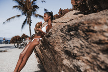 Serious Young Hottie Black Female Is Drinking Fresh Coco Water While Leaning On The Cliff With Beach And Palms In The Background; Sexy Brazilian Girl In Swimsuit Is Slaking Her Thirst Near The Rock