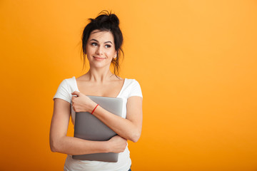 Portrait of fascinating woman with trendy hairstyle hugging modern silver laptop and looking aside with brooding sight, over yellow wall copy space