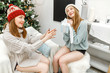 © EdNurg - happy girl friends exchanging presents in christmas decorated living room in funny knitted hats