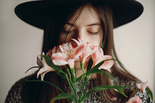 Stylish Hipster Girl In Hat Holding Pink Flowers And Petals On Sweater Growing. Boho Woman With Beautiful Alstroemeria In Hands, Smelling Flower. Creative Sensual Female Portrait. Calm Face