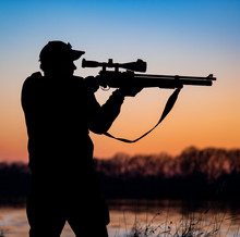 Silhouette Of Hunter With Rifle On The Background Of Sunset. Man With A Gun Against The Evening Sky And The River