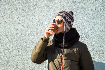 Wall Mural - Young man drinks coffee to go from paper cup. Handsome teenage male person in sunglasses and knitted hat drinking hot beverage on bright sunny winter day