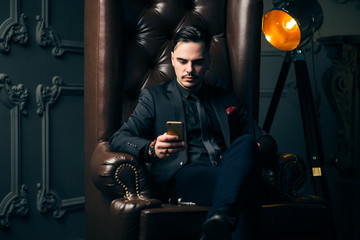 Young handsome man posing for a fashion shoot in a studio. Fashion as a lifestyle. Man wearing a beard. Model sitting on a sofa in tuxedo. Successful fashionable businessman. Business look advertising