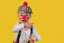 Child Clown Smile Shows A Finger Copy Space Area On Yellow Background. Funny Little Clown With Red Nose. Concept Birthday, Day 1 April, Party. Beautiful Portrait Kid Jester In Studio.