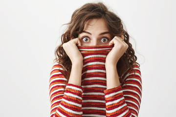 Maybe no one will notice me like this. Portrait of funny cute curly-haired female hiding in sweater and staring at camera with shocked or surprised expression, standing over gray background