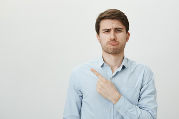 Such shame I missed all fun. Portrait of handsome sad caucasian guy pointing left with index finger whining and looking gloomy, being upset with issue or jealous, standing over gray background