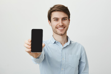 If you seek new phone, buy this model. Portrait of cheerful attractive white male with bristle, pulling hand towards camera and showing smartphone, smiling broadly over gray background