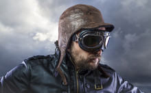 Old Plane Pilot On Background Of Storm Clouds With Expressive Face. Glasses And Old Hat With Black Leather Jacket