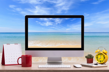 Fototapete - Modern desktop computer, Coffee cup, alarm clock, notebook and calendar on wooden table and view of tropical beach. Saved with clipping path. Work and holiday concept