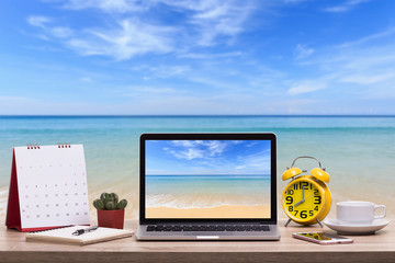 Fototapete - Modern laptop computer, Coffee cup, alarm clock, notebook and calendar on wooden table and view of tropical beach. Saved with clipping path. Work and holiday concept