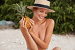 © WHstudio Leushin N - Beautiful happy young female with nude body, wears straw hat on head, sits on beach alone, keeps pineapple in hand, smiles happily and looks down likes exotic fruits. People, summer, nutrition concept