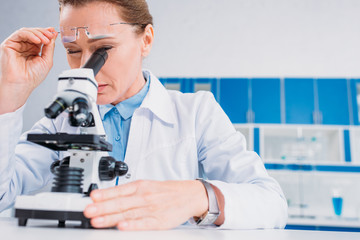 Wall Mural - female scientist in lab coat and eyeglasses looking through microscope on reagent in lab