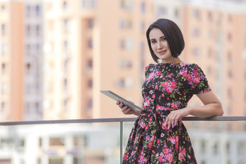 Smart beautiful caucasian fit business woman with a strong face type and dark bob short hairstyle in casual romantic dress in european city streets with tablet in hands, working, smiling. copy space