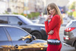 Beautiful rich luxury looking blonde caucasian business woman in smart-casual red outfit standing on a european streets near cars, holding tablet, distant working. Daylight, outdoors.