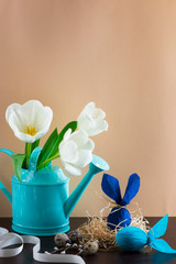 Blue watering can with white tulips and two blue Easter eggs in form of rabbit on brown background.