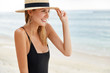 © WHstudio Leushin N - Adorable cheerful woman wears black bikini and straw hat, looks positively into distance while stands against ocean view, admires endless horizon. Relaxed female strolls on beach outdoor near sea