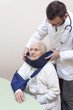 Staruszka w białym szlafroku siedzi na krześle z temblakiem na ręce. Lekarz zakłada jej na szyję kołnierz ortopedyc zny. 