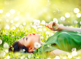 Beautiful young woman lying on the field in green grass and blowing dandelion flowers. Allergy free concept