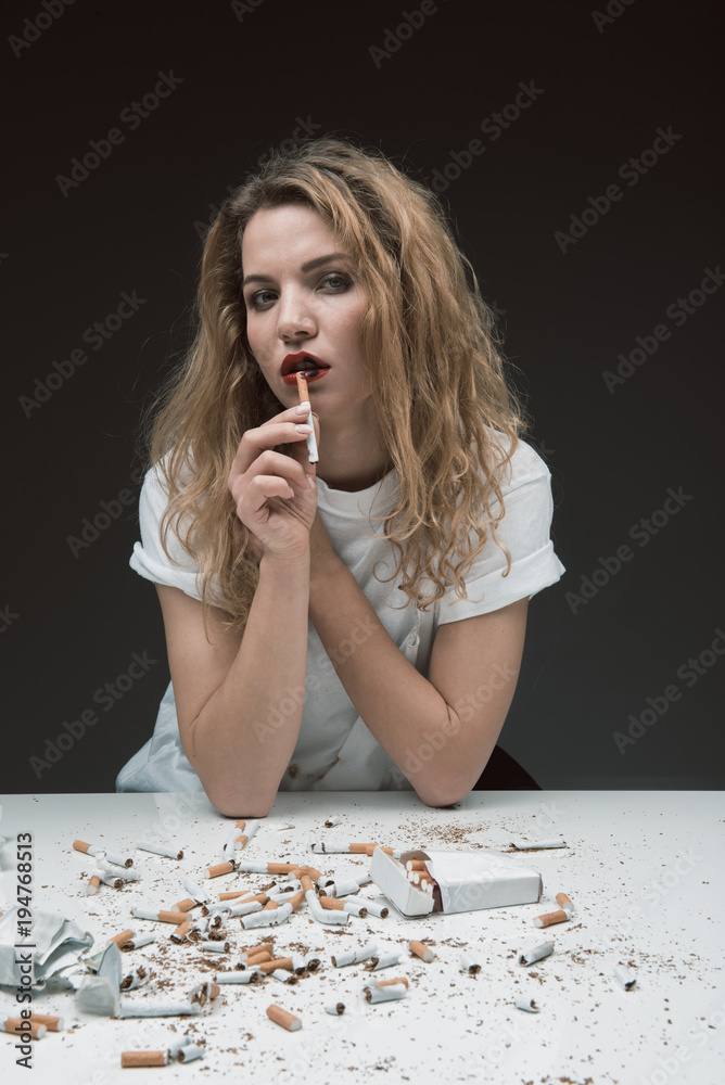 eating a cigarette Portrait of unhealthy serene girl holding cigarette near her mouth and  looking at camera, cigarettes are scattered all over the table in front of  her. Isolated on background Stock Photo