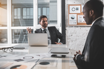 Wall Mural - Portrait of grave boss with beard in suit is sitting at desk with computer while looking at his employee discontentedly. African manager is feeling upset