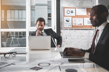 Wall Mural - Steely glance. Portrait of serious young bearded man in suit is sitting at table with laptop and looking at camera confidently. Profile of african manager is typing on computer with concentration