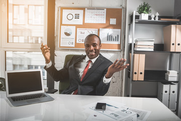 Poster - Smart entrepreneur. Portrait of joyful confident african businessman in formal wear is sitting at table with open laptop and smartphone while looking at camera with smile. He is gesturing positively
