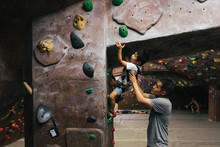 Little Boy Bouldering In Indoor Climbing Gym With Father's Help