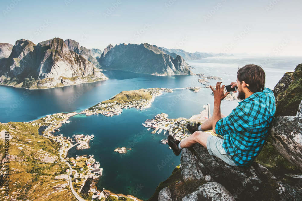 Man tourist using smartphone on cliff Travel lifestyle concept adventure outdoor in Norway Reinebringen mountains aerial view Lofoten islands summer vacations - obrazy, fototapety, plakaty 
