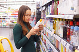 Happy young brunette selecting bottle of child's shampoo in beauty store.