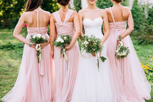 Three Bridesmaids In Powdery Dresses Transformers With Bouquets In Hands Stand With Their Backs Near The Bride In A White Dress With A Wedding Bouquet In Her Hand On A Green Lawn