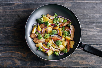 Poster - Fried potatoes with meat, broccoli and red onions decorated with rosemary in a frying pan on a wooden table top view